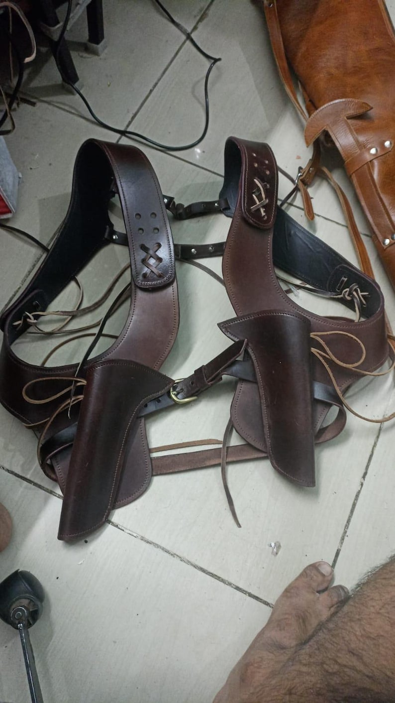 May include: A pair of brown leather gun holsters with attached shoulder straps and belts. The holsters are designed to be worn across the chest, with intricate stitching and decorative details. The leather appears to be high-quality, with a rich, dark brown color.