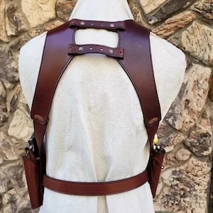 May include: A brown leather gun holster with two attached pouches and a belt. The holster has straps that go over the shoulders and connect at the back. The leather is a rich, dark brown color.