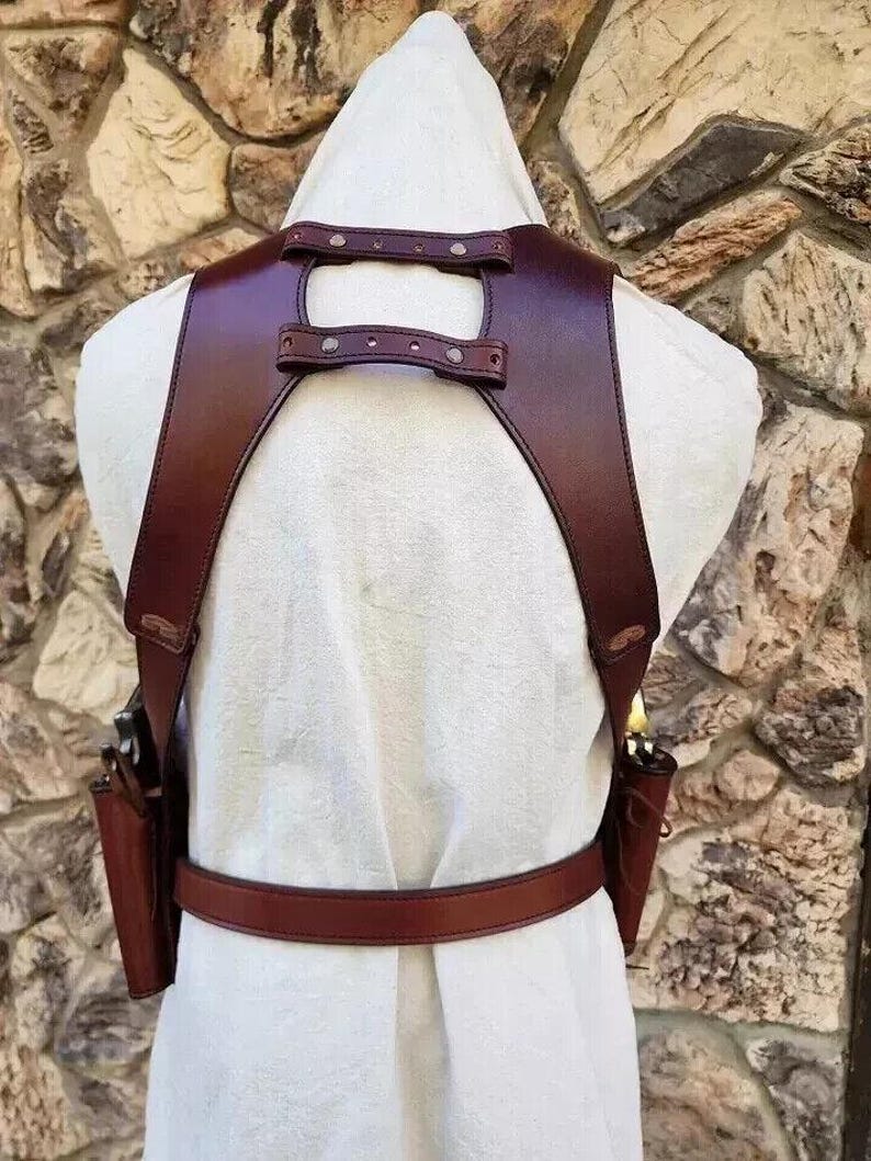 May include: A brown leather gun holster with two attached pouches and a belt. The holster has straps that go over the shoulders and connect at the back. The leather is a rich, dark brown color.