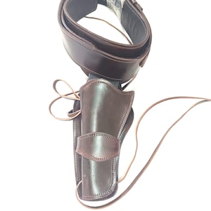 Op de afbeelding: Een bruine leren holster- en riemset. De holster is ontworpen om een ​​handvuurwapen vast te houden en is bevestigd aan een brede riem. De riem is donkerbruin en heeft een gesp. Een leren riem is aan de holster bevestigd.