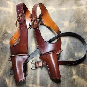 May include: A brown leather shoulder holster with two gun holsters and a belt. The holster has a two-tone design, with lighter brown accents. The leather is stitched and has decorative details. The belt has a silver buckle.