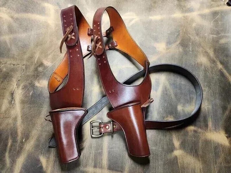 May include: A brown leather shoulder holster with two gun holsters and a belt. The holster has a two-tone design, with lighter brown accents. The leather is stitched and has decorative details. The belt has a silver buckle.