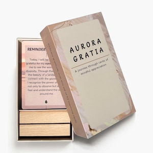 May include: A set of "AURORA GRATIA" mindful appreciation cards in a decorative box. The box and cards have a neutral color palette with the text "AURORA GRATIA" and "A journey through cards of mindful appreciation". The open box reveals a card with the text "REMINDER".