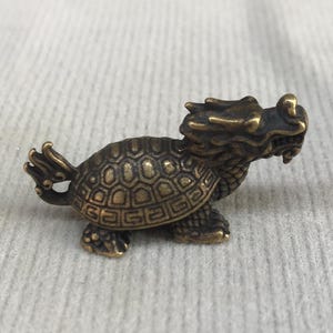 May include: A small, antique-style brass figurine depicting a mythical creature with the head of a dragon and the body of a turtle. The sculpture features detailed scales on the shell, a textured dragon head, and a small tail, finished in a dark bronze tone.