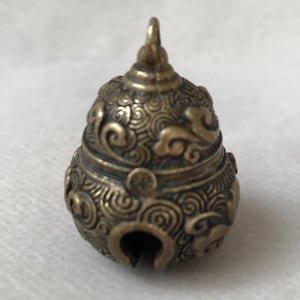 May include: A small, pear-shaped brass bell featuring an intricate, embossed swirling pattern across its entire surface. The bell has a small loop at the top for hanging and a single vertical opening at the bottom, showcasing a metallic finish.