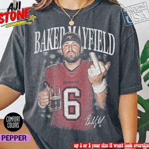 May include: A charcoal grey short-sleeve t-shirt featuring a graphic of a football player in a red jersey with the number 6 and the text BAKER MAYFIELD. The player is holding a beer mug and gesturing with a middle finger. The shirt has a vintage wash.