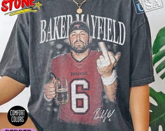 Puede incluir: Una camiseta gris carbón de manga corta con un gráfico de un jugador de fútbol americano con una camiseta roja con el número 6 y el texto BAKER MAYFIELD. El jugador sostiene una jarra de cerveza y hace un gesto con el dedo medio.