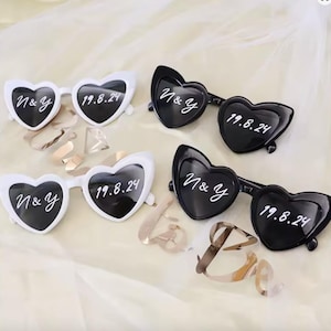 Puede incluir: Gafas de sol en forma de corazón en blanco y negro. Las gafas tienen el texto "N&Y" y la fecha "19.8.24" impresos en las lentes. La imagen muestra cuatro pares de gafas de sol, perfectas para una boda o fiesta.