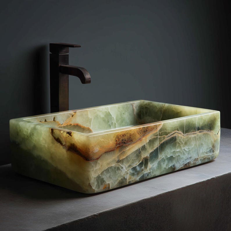 May include: A rectangular, green onyx bathroom sink with brown and yellow veining. The sink is paired with a dark bronze faucet. The sink is set against a dark gray wall and a gray countertop.