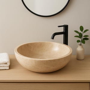 Op de afbeelding: Een beige, ronde wastafel met een getextureerd oppervlak staat op een lichte houten wastafel. Een zwarte kraan en een kleine vaas met groen staan rechts. Een zwart omlijste spiegel hangt boven de wastafel. Een opgevouwen witte handdoek staat links.