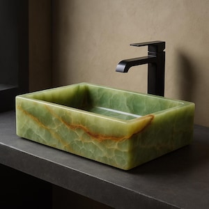 May include: Rectangular green onyx bathroom sink with brown veining. The sink is paired with a matte black faucet. The sink is made of a solid block of stone and sits on a dark gray countertop.