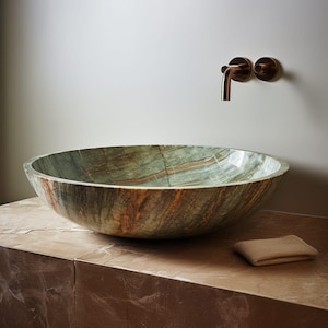 May include: A large, oval-shaped stone sink with green and brown veining. The sink is set on a brown countertop, with a bronze faucet mounted on the wall behind it. A small, tan washcloth rests on the counter.