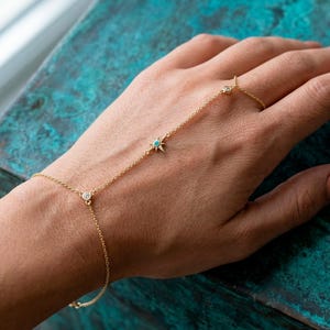 Puede incluir: Una delicada pulsera de cadena de mano dorada con un dije de estrella con una piedra turquesa, una piedra redonda transparente y un anillo. La pulsera está conectada por una fina cadena. La joya se muestra en una mano.