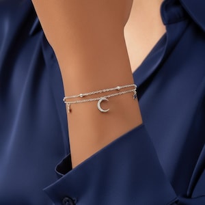 Pode incluir: Uma pulseira de prata de duas voltas com berloques celestiais. A pulseira apresenta um berloque de lua crescente com pequenos cristais, um berloque de estrela e pequenos berloques pendurados. A pulseira é usada em um pulso contra um fundo azul escuro.