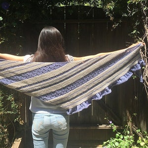 May include: A triangular knit shawl featuring alternating diagonal stripes of light beige and periwinkle blue yarn. The design incorporates sections of openwork eyelet stitches and a ruffled edge along the bottom border of the garment.