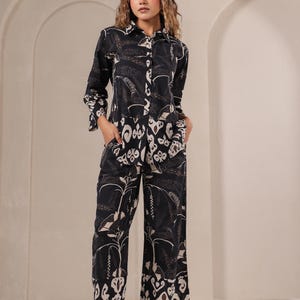 May include: Black button-down shirt and wide-leg pants set with a white and brown floral pattern. The long-sleeved shirt has a collared neckline and pockets. The pants have a matching print. The outfit is made of a soft, flowing fabric.