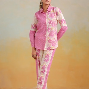 May include: A two-piece floral print outfit in pink and cream. The long-sleeved button-down shirt has a pink patterned side and a cream floral side. The matching pants have a floral border. The outfit is worn with white sandals.