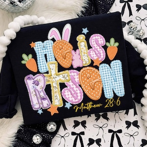 May include: Black long-sleeve shirt with the words "He is Risen" in colorful, appliqued letters. The design includes Easter-themed elements like carrots, bunny ears, and a cross. The text "Matthew 28:6" is written in a script font.