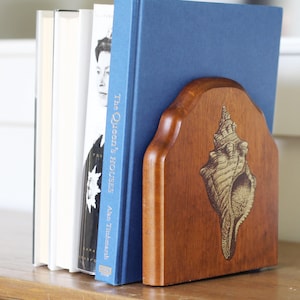 May include: A wooden bookend with a detailed seashell design, holding several books. The bookend is a warm brown color, and the books have white, blue, and black covers. The bookend is on a wooden surface.