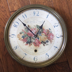 May include: Round wall clock with a gold-colored metal frame. The clock face features a floral bouquet with pink and yellow roses, blue numerals, and the text "Le Jardin d'Emilie" and "Fleuriste".