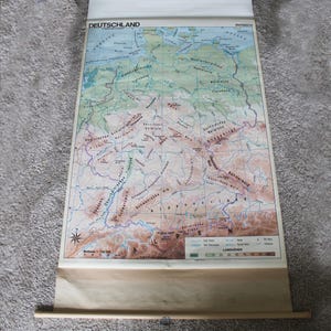 May include: A vintage map of Germany, titled "DEUTSCHLAND," with detailed geographical features. The map is rolled up and has a light brown wooden dowel at the bottom. The map's colors include shades of green, brown, and blue.
