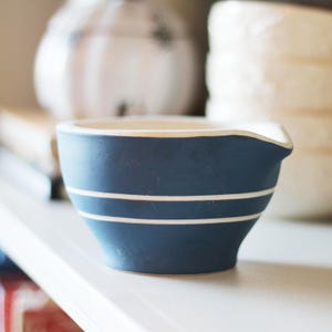 May include: A small, blue ceramic bowl with two white stripes and a pouring spout. The bowl has a cream-colored interior and is likely used for mixing or serving. Other white ceramic items are in the background.