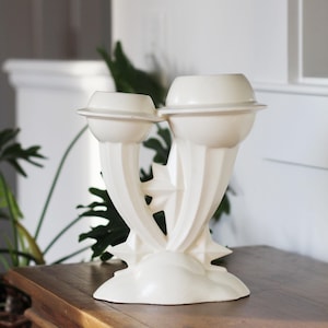 May include: White ceramic vase with a unique, abstract design. The vase has two openings at the top and a star-shaped base.