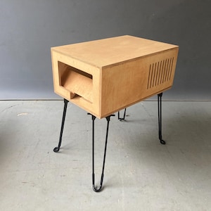 May include: A light-coloured wooden side table with a rectangular body and a cut-out section on the front. The table has black hairpin legs and vertical slits on one side.
