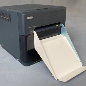 Peut inclure: Une imprimante photo numérique DNP gris foncé avec un bac de sortie papier blanc. L'imprimante porte le logo DNP et des informations sur le modèle à l'avant. L'imprimante est conçue pour l'impression photo.