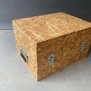 Op de afbeelding: Een grote, rechthoekige houten kist van spaanplaat. De kist heeft een natuurlijke houtnerf en is vastgezet met metalen grendels en een handvat. De kist staat op een grijze ondergrond.