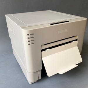 Peut inclure: Une imprimante beige avec un bac de sortie papier ouvert. L'imprimante comporte des voyants lumineux indiquant "POWER", "RIBBON", "PAPER" et "ERROR". L'imprimante est posée sur une surface grise.