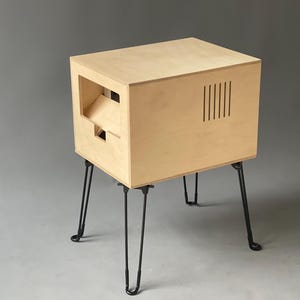 Peut inclure: Une table d'appoint en bois clair de forme carrée. Elle présente une découpe sur un côté et des fentes verticales sur un autre. La table est soutenue par quatre pieds en épingle à cheveux noirs, créant une esthétique moderne.