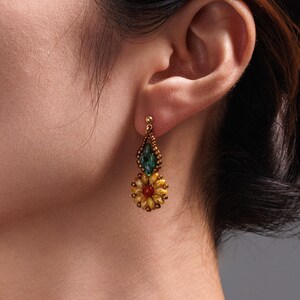 May include: Gold-coloured drop earrings with a floral design. The earrings feature a teardrop-shaped green stone above a flower with yellow petals and a red centre. The earrings are attached to the ear with a small gold stud.