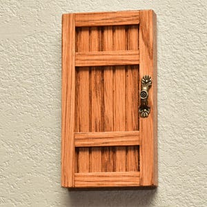 May include: A rectangular wooden door with a natural light brown wood grain finish, featuring vertical slats and two horizontal crossbars. A small, ornate, antique brass-colored metal handle is attached to the right side of the door frame.