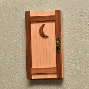 May include: Wooden decorative door with a crescent moon cutout and a decorative handle. The door is made of light-colored wood with darker wood trim. The handle is a bronze color.