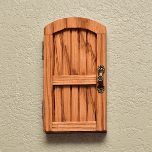 May include: A small, arched wooden door with a light brown wood grain finish, featuring vertical planks, a horizontal crossbar, and an ornate, antique-style brass-colored metal handle attached to the right side, mounted on a textured wall.