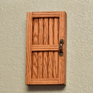 May include: A miniature wooden door with a detailed, vertical-slat design. The door is stained a warm brown and features a decorative, antique-style handle. The door is mounted on a textured, light-colored wall.
