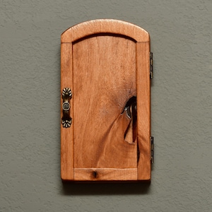 May include: A small, wooden cabinet with a natural finish. The cabinet has a decorative handle and hinges. The wood grain is visible, and there is a unique knot detail on the door. The cabinet is mounted on a gray wall.