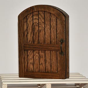 May include: A small, arched wooden door featuring a dark brown wood grain finish with vertical planks and a horizontal crossbar. The door is equipped with a decorative, antique-style brass handle attached to the right side of the door frame.
