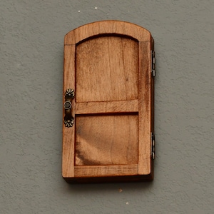 May include: A small, wooden door with a curved top and a rectangular lower section. The door is a warm brown color with visible wood grain. It features decorative metal hinges and a handle. The door is mounted on a gray wall.