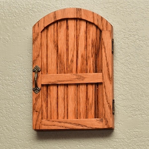 May include: A wooden cabinet door with a rounded top and vertical plank design. The door is stained a warm reddish-brown color and features a decorative brass handle and hinges. The cabinet is mounted on a textured, light-colored wall.