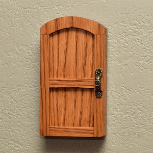May include: A small, arched wooden door with a natural oak grain finish and a horizontal crossbar. The door features a decorative, antique-style brass handle with floral detailing mounted on the right side, providing a classic look.