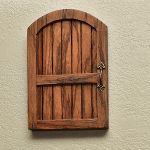 May include: A small, wooden door with a rounded top and vertical planks. The door is stained a warm brown with darker graining. It features a horizontal brace and an ornate, brass-colored handle. The door is mounted on a textured, light-colored wall.