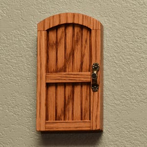 May include: A small, arched wooden door with a natural light brown wood grain finish. The door features vertical planks, a horizontal crossbar, and a decorative, antique-style brass-colored metal handle attached to the right side.