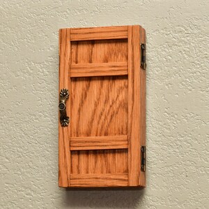 May include: A rectangular wooden wall cabinet with a natural oak finish, featuring a single door with two horizontal crossbars. The door is equipped with a decorative antique brass-toned metal handle and is attached with two matching metal hinges.