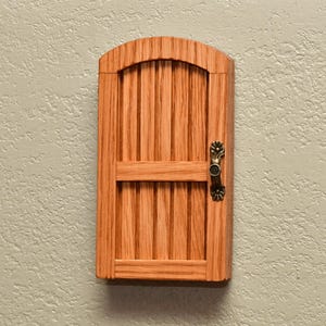 May include: A small, arched wooden door featuring vertical slats and a horizontal crossbar. The door is crafted from light brown wood with a visible grain pattern and is equipped with a decorative, antique-style brass metal handle on the right side.