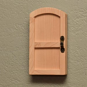 May include: A small, arched wooden door made of light-colored natural wood with a visible grain. The door features a horizontal crossbar and a decorative, antique-style brass-toned metal handle attached to the right side of the door frame.