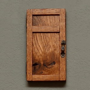 May include: A small, rectangular wooden cabinet with a door. The cabinet is made of light brown wood with a visible wood grain. It has a decorative handle and hinges. The cabinet is mounted on a gray wall.