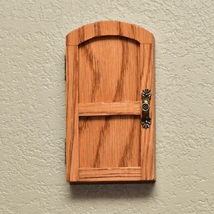 May include: A small, arched wooden door with a light brown wood grain finish. The door features a horizontal crossbar and a decorative, antique-style brass-colored metal handle on the right side. Two small metal hinges are visible on the left side.