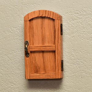 May include: A small, arched wooden door with a natural wood grain finish. The door features a horizontal crossbar, a decorative antique brass-colored metal handle on the left side, and two matching metal hinges attached to the right side frame.